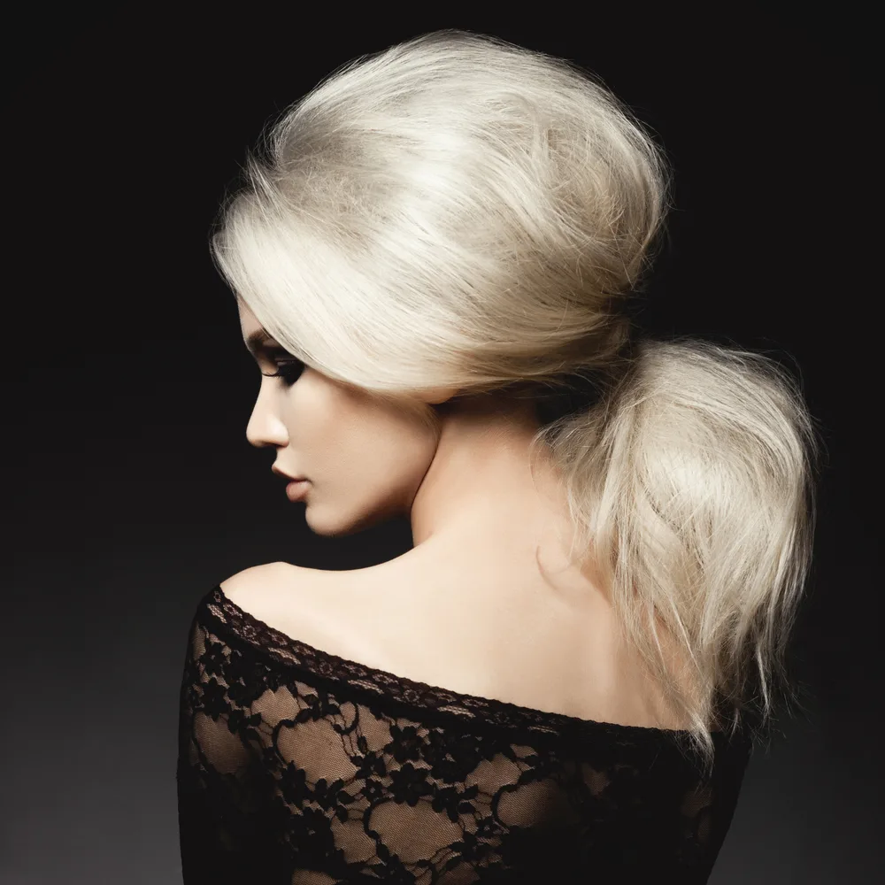 Rear view of a blonde woman turning to the side in a black lacy off-shoulder top to show off one of the more upscale teased low ponytail hairstyles