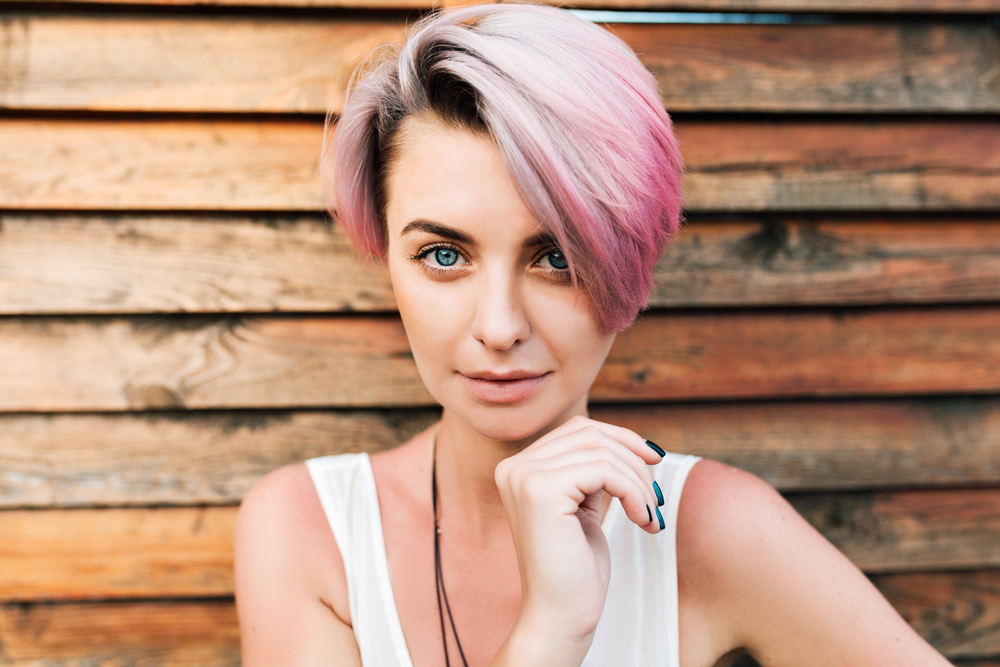 Woman in front of a wooden plank wall with hand under her chin shows off pink hair styled in one of the top fine hair thin hair low maintenance short hairstyles listed in this article