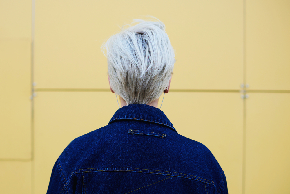 Back view of a woman with platinum blonde pixie cut wearing a messy hairstyle in front of a yellow wall