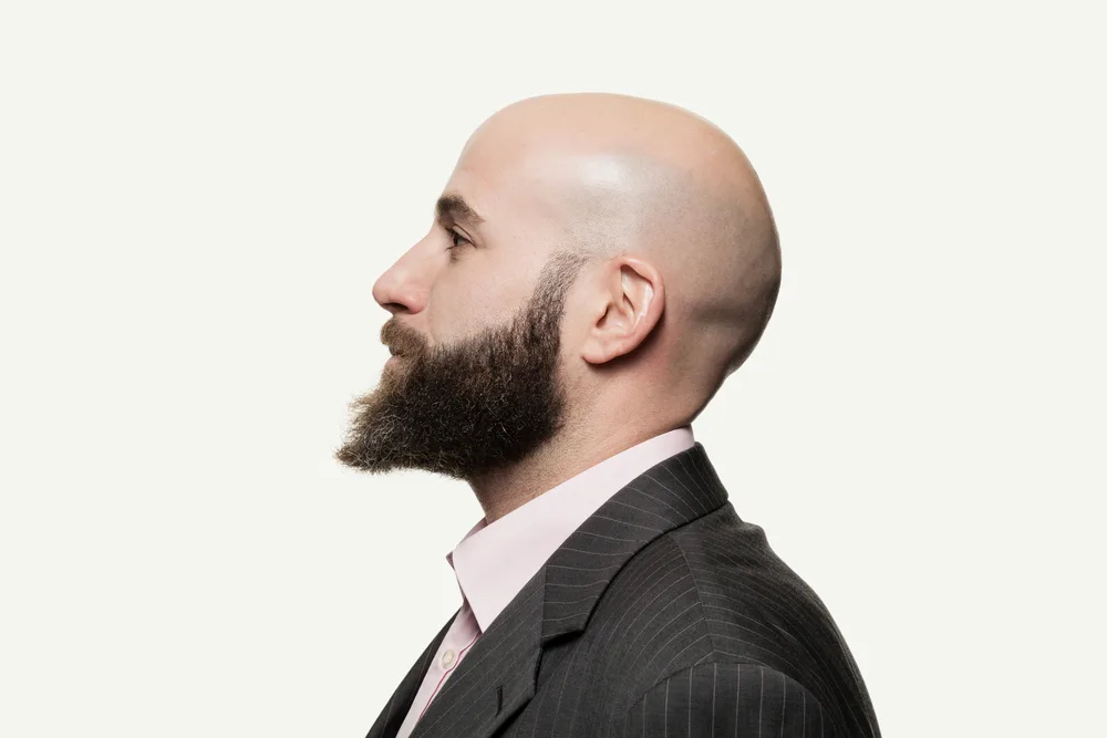 Side view of bearded man with bald head rocking one of the top low maintenance haircuts for men with a suit and tie on