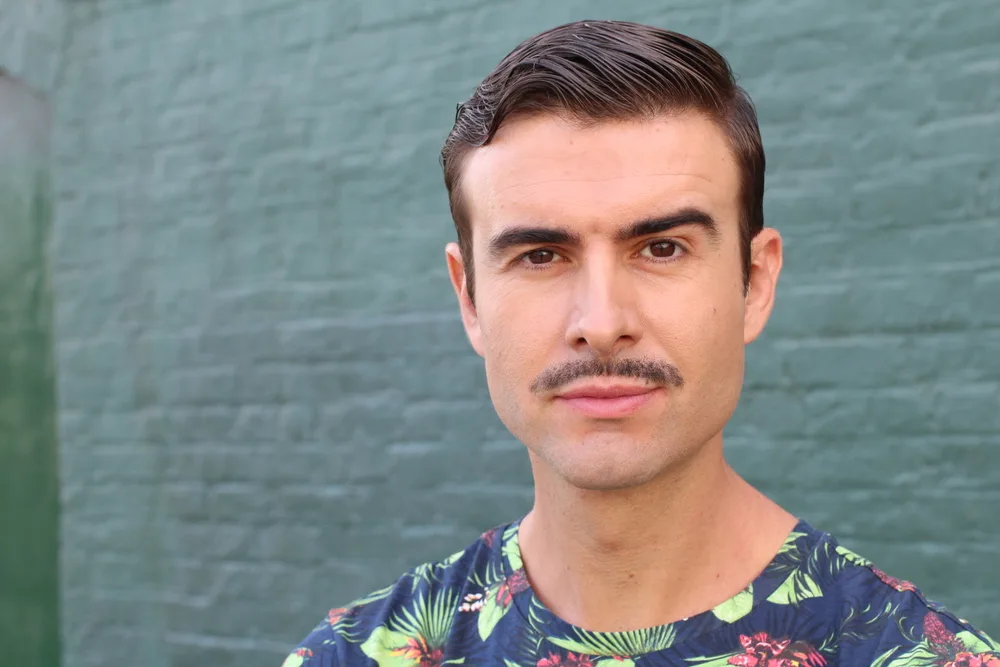 Brunette man with a patterned shirt stands in front of a painted brick wall wearing one of the most well-known mustache styles, the pencil mustache