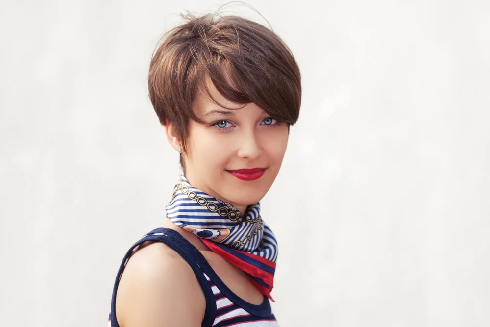 Brunette woman wearing a scarf and striped tank top smiles with a mushroom haircut and bangs, listed as one of the best fine hair thin hair low maintenance short hairstyles 