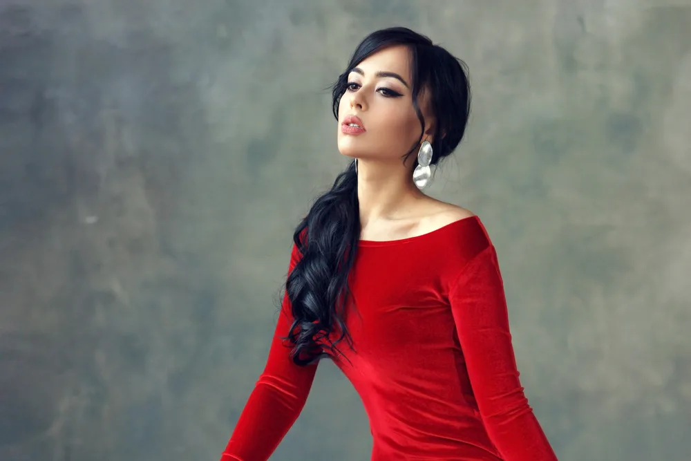 Dark-haired woman in red long-sleeved dress looks away with head lifted to show what glamorous ponytail hairstyles look like with curled ends