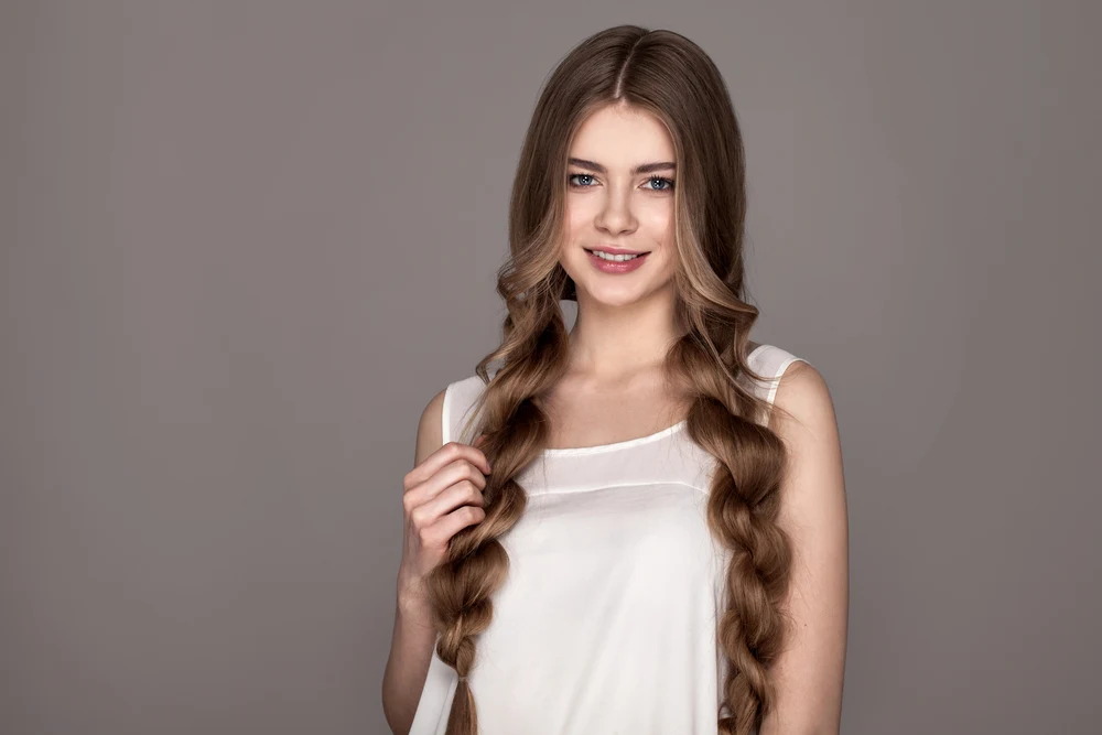 Woman in white sleeveless top models long rope braids and smiles in front of a gray wall