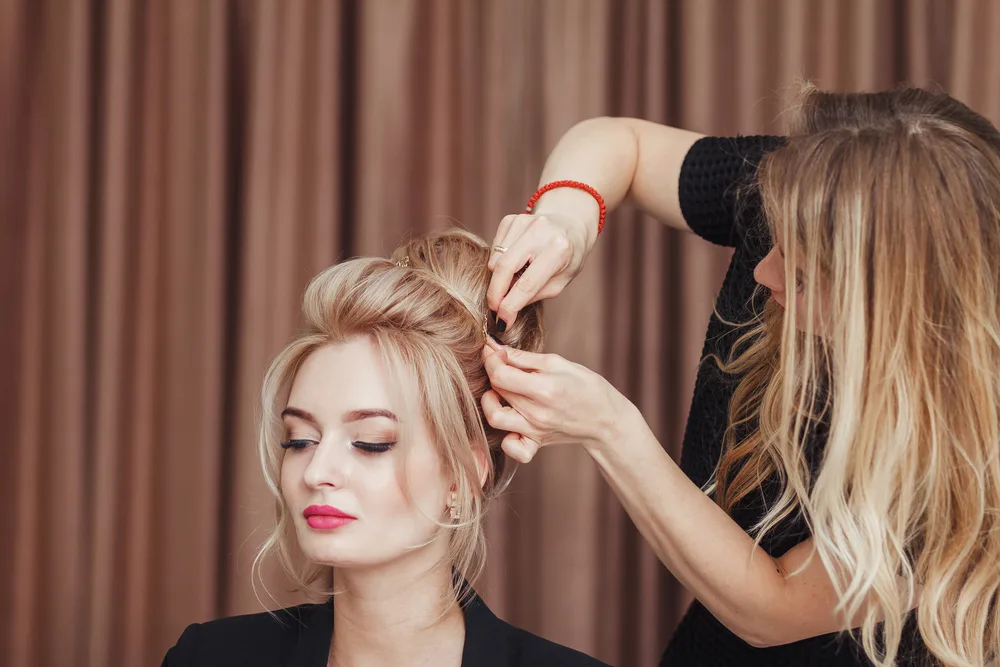 Stylist finishes a blonde woman's formal hairstyle with bobby pins to show examples of cute homecoming hairstyles