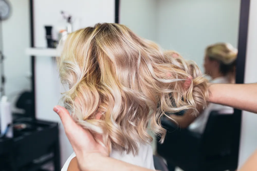 Hair stylist lifts finished curly blonde hair after giving her one of the best low maintenance medium length hairstyles for thick hair
