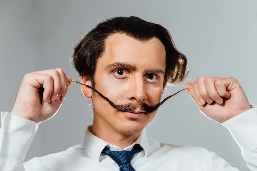 An eccentric hipster man holds the ends of his Dali mustache, one of the most unique mustache styles with upward tilt and length that extends far past the mouth 